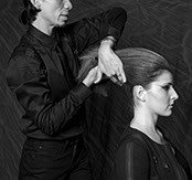 Black-and-white portrait of a stylist shaping a client's hair into an updo, both shown in profile