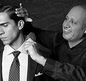 Black-and-white portrait of a male stylist brushing the shoulder of a client's slicked-back hairstyle